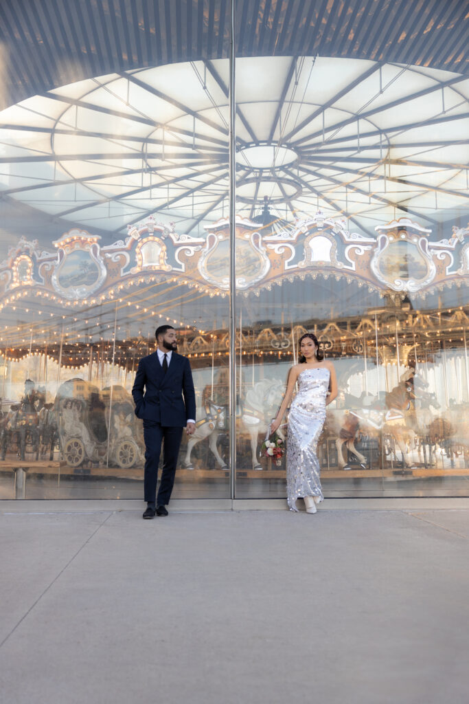 Couple standing in front of Jane's Carousel in Brooklyn Bridge Park, with its intricate lights and reflections creating a whimsical and romantic scene.