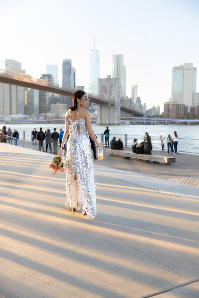 Bride walking along the Brooklyn waterfront, holding a bouquet with the Manhattan skyline and Brooklyn Bridge beautifully framed in the background.