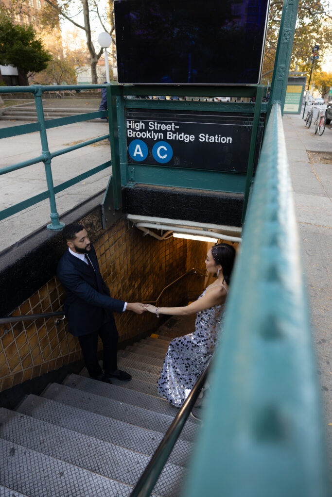 Couple holding hands at the entrance of the High Street - Brooklyn Bridge Station, framed by the green railing and city surroundings.